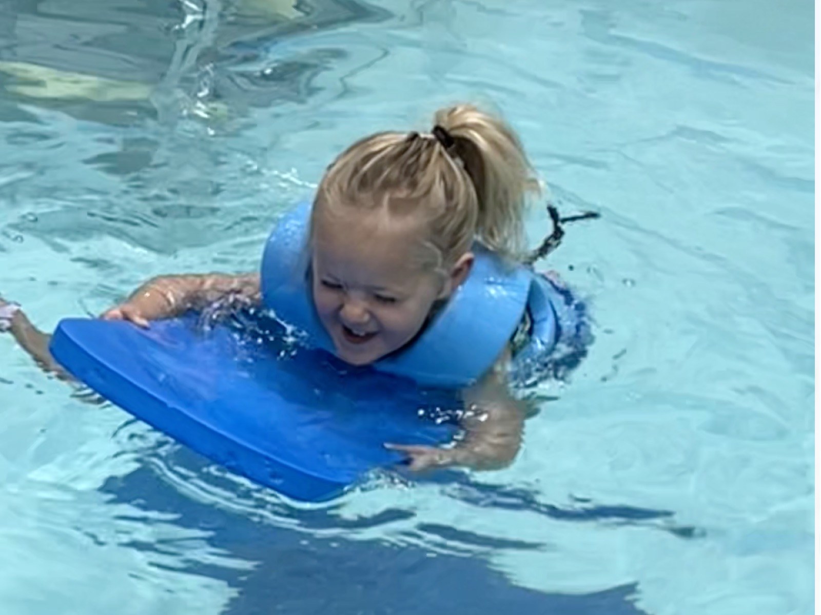 My daughter laughing as she paddles across a pool holding a foam kickboard